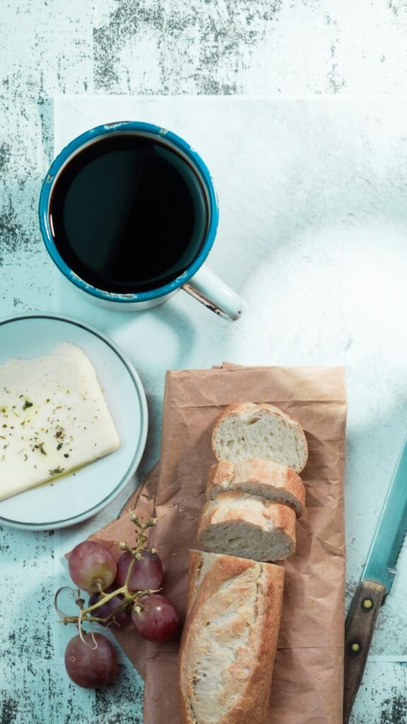 Top-down view of a rustic breakfast table with black coffee, sliced bread, cheese, and grapes.