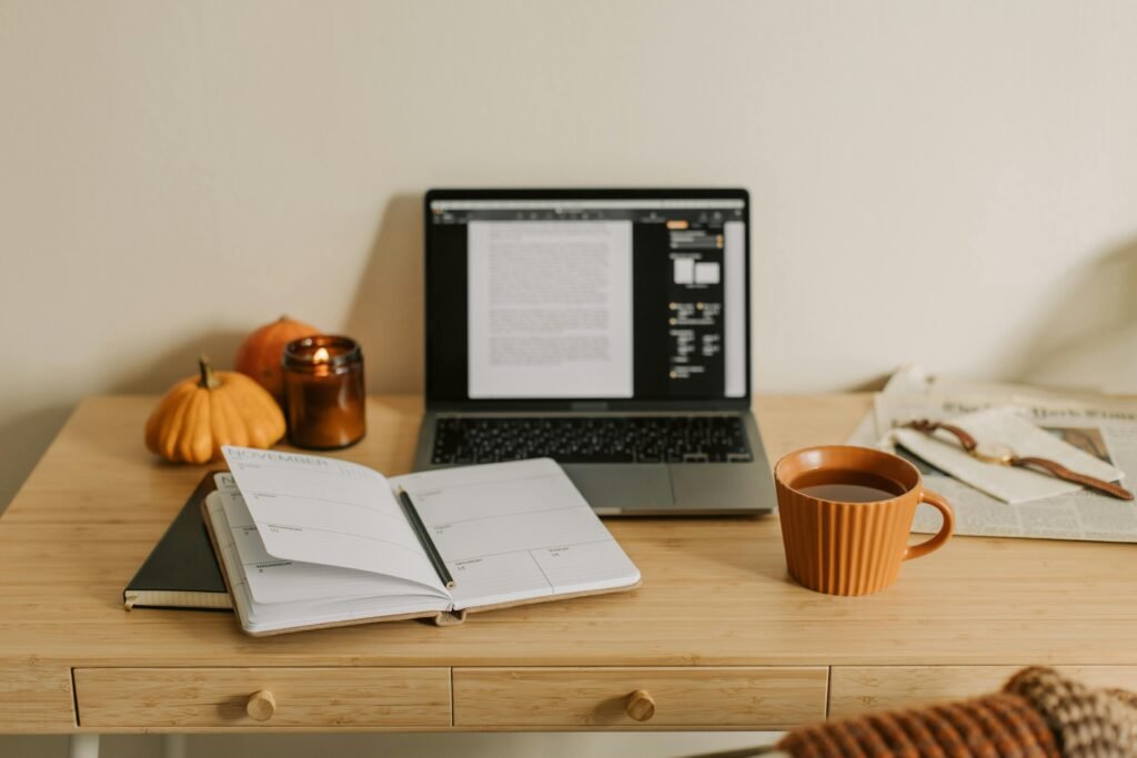 A cozy workspace featuring a laptop, coffee, notebook, book, homeschool planner and pumpkins in autumn setting.